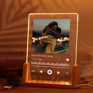 Op de afbeelding: Een gepersonaliseerd nachtlampje met een foto van twee mensen die elkaar omhelzen. Het heeft een houten voet en een helder acryl paneel met de tekst "Your Favourite Song" en muziekbedieningspictogrammen.