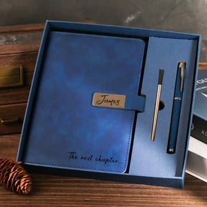 Peut inclure: Un ensemble cadeau comprenant un journal en cuir bleu et un stylo. Le journal porte une plaque signalétique métallique avec le nom "James" et le texte "The next chapter...". L'ensemble comprend un stylo et un stylet.