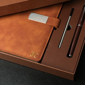 Puede incluir: Un diario de cuero marrón con un cierre de metal plateado y las iniciales "JC" grabadas en dorado. El diario se presenta en una caja marrón con un bolígrafo plateado y un bolígrafo marrón oscuro.