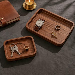 May include: Three wooden trays with personal items. The largest tray has a watch, ring, and a pin. The middle tray has a watch and a ring. The smallest tray has keys and a ring. The trays have engraved text. A leather notebook and a lit candle are also present.