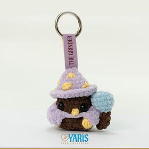 May include: A handmade crochet keychain featuring a brown creature wearing a purple wizard hat with yellow stars and a matching scarf. The keychain has a blue pom-pom and a tag that reads "THE GRINDER". The keychain is attached to a silver ring.