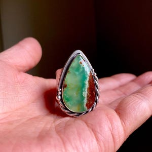 May include: A silver ring with a large, teardrop-shaped stone. The stone has green and brown colours. The ring has a twisted rope design on the sides and small silver beads near the top.
