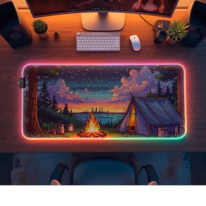 May include: A rectangular mouse mat with a pixelated camping scene, including a tent, campfire, and starry night sky. The mat has a glowing, multi-coloured edge. Computer accessories surround the mat on a wooden desk.