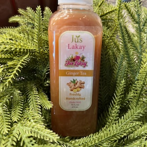 Peut inclure: Une bouteille en plastique transparente remplie de th&eacute; au gingembre, &eacute;tiquet&eacute;e "Jus Lakay". L'&eacute;tiquette indique "Ginger Tea" et "Freshly Handcrafted". La bouteille est entour&eacute;e de feuillage vert. Contient 473 ml de th&eacute;.