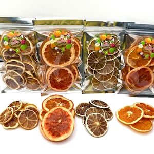 Puede incluir: Variedad de rodajas de frutas cítricas deshidratadas, incluyendo naranjas, limones y limas. Algunas rodajas están empaquetadas en bolsas de plástico transparente con etiquetas coloridas. Las rodajas de fruta están dispuestas sobre una superficie blanca.