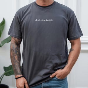 Könnte beinhalten: Dunkelgraues T-Shirt mit dem weißen Text "duck fan for life". Das Shirt ist einfarbig und hat einen Rundhalsausschnitt. Die Person, die das Shirt trägt, hat Tätowierungen auf dem Arm und trägt ein schwarzes Armband.