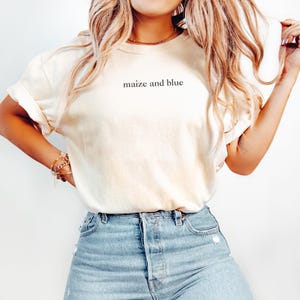 Peut inclure: T-shirt crème avec l'inscription "maize and blue" en noir. Le t-shirt est associé à un jean en denim bleu clair. L'ensemble dégage une ambiance décontractée et simple, idéal pour un look quotidien.