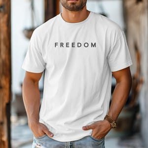 May include: White t-shirt with the word "FREEDOM" printed in dark grey. The shirt has short sleeves and a crew neck. The model is wearing a watch with a brown strap.