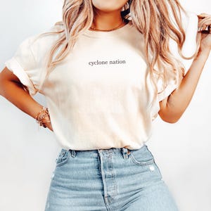 Può includere: Una t-shirt a maniche corte color crema con la scritta "cyclone nation" stampata in piccole lettere minuscole nere al centro del petto. La maglietta presenta un girocollo ed è abbinata a un paio di jeans in denim azzurro.