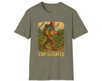 May include: Olive green t-shirt featuring a Bigfoot illustration with a backpack and walking stick, set against a mountain and forest backdrop. The text "STAY ELEVATED" is printed below the graphic. A casual tee for outdoor enthusiasts.