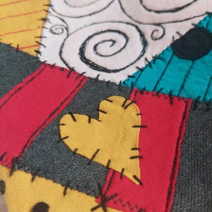 May include: A fabric patchwork textile featuring a variety of colors including yellow, red, teal, and grey. The design includes a yellow heart shape, black stitched detailing along the seams, and white fabric sections with black spiral patterns.