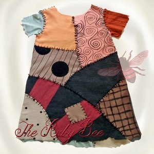 May include: A short-sleeved dress featuring a patchwork design with various fabric patterns including black polka dots, swirls, stripes, and solid panels in peach, red, and dark gray, joined by visible black stitching. The text The Ruby Bee is present.