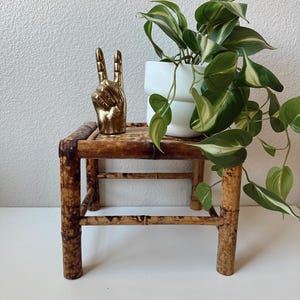 May include: A gold-colored hand statue displaying the peace sign sits on a small, square bamboo table. A white ceramic pot with a green and white plant is on the table. The table has a natural, brown finish.