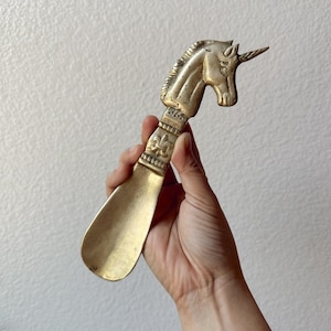 May include: A brass shoe horn with a unicorn head design. The horn features a long, curved blade and a handle with a detailed unicorn head and a fleur-de-lis pattern. The metal has a warm, antique finish.