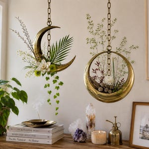 May include: Two hanging brass planters suspended by chains. One is crescent moon-shaped, filled with white flowers, green palm leaves, and trailing ivy. The other is a circular ring shape containing small branches, white flowers, and mossy textures.