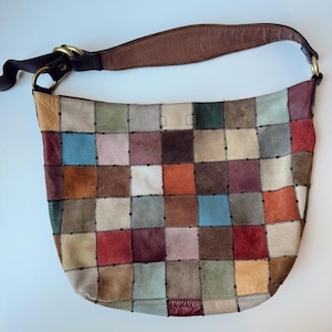 May include: A hobo-style shoulder bag constructed from a patchwork of multi-colored leather squares in shades of red, blue, green, brown, and cream. The bag features a single dark brown leather strap attached with brass-toned metal rings.