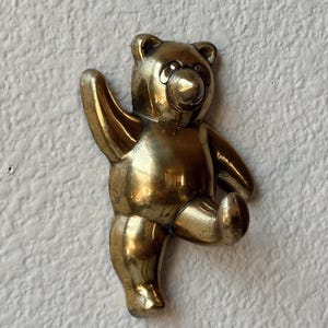 May include: A polished brass wall hook shaped like a teddy bear. The bear is depicted in a standing position with one arm raised upward and one leg bent forward, creating a curved hook shape for hanging items on its arm and leg.