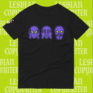 Peut inclure: T-shirt noir avec trois têtes d'extraterrestres violets dans les poses « ne rien voir, ne rien entendre, ne rien dire ». Les extraterrestres ont de grands yeux noirs. Le texte "LESBIAN COPYWRITER" est imprimé en arrière-plan.