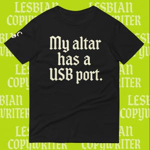 May include: Black t-shirt featuring the phrase "My altar has a USB port" in a gothic font. The shirt has a crew neck and short sleeves. The background has the words "LESBIAN COPYWRITER" in a repeating pattern.