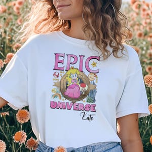 May include: White t-shirt featuring a cartoon graphic of Princess Peach from the Super Mario franchise. The design includes the text "EPIC UNIVERSE Kate" in pink, with additional cartoon elements like mushrooms and a rainbow.