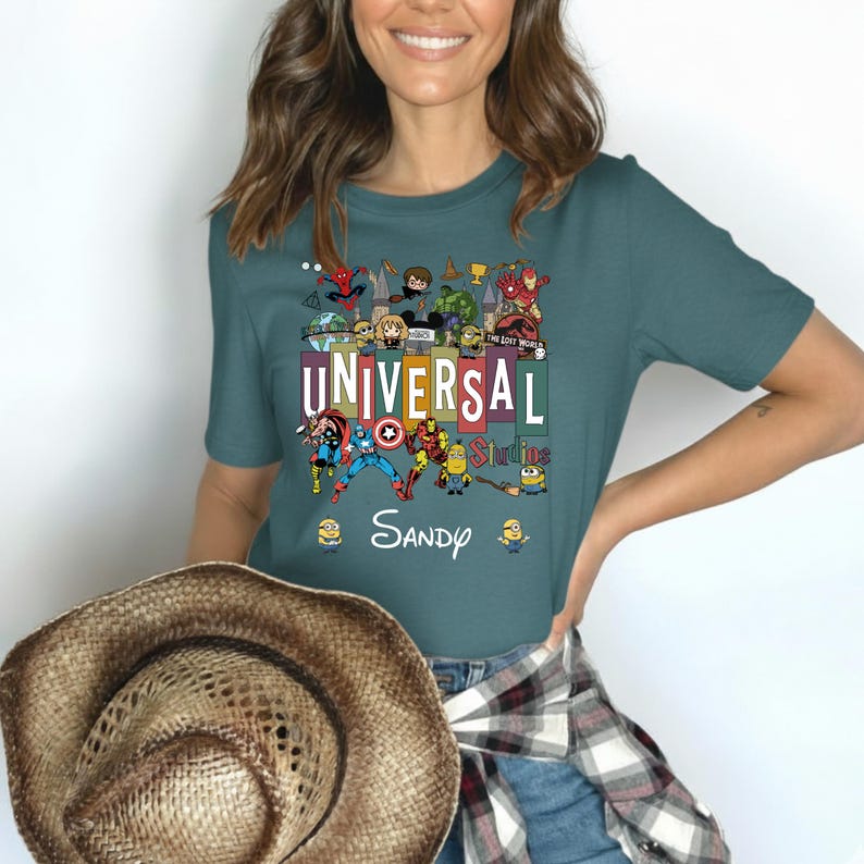 Op de afbeelding: Tealkleurig T-shirt met een kleurrijke afbeelding van Universal Studios-personages, waaronder Spider-Man, Captain America en Minions. De tekst "UNIVERSAL Studios" en de naam "Sandy" zijn ook zichtbaar. Een bruine hoed wordt in de afbeelding vastgehouden.