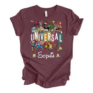 Op de afbeelding: Bordeaux t-shirt met een kleurrijke afbeelding van Universal Studios-personages, waaronder Spider-Man, Minions en superhelden. De tekst "UNIVERSAL Studios" staat prominent afgebeeld, met de naam "Sophia" eronder. Een leuke, thematische tee.