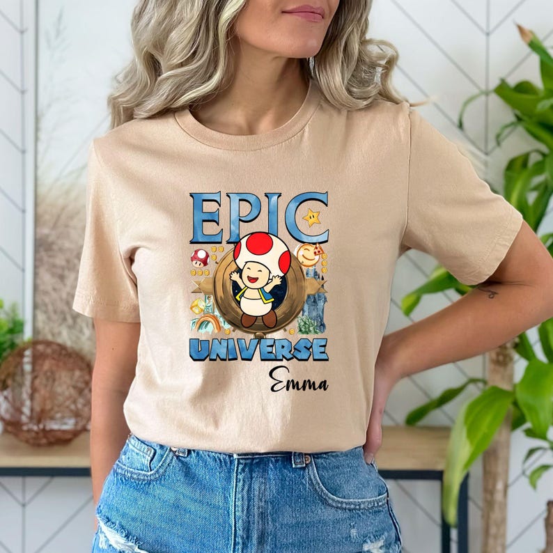 May include: Beige t-shirt featuring the text "EPIC UNIVERSE Emma" in blue and black. The graphic includes a cartoon mushroom character, a rainbow, and other colorful elements. The shirt has short sleeves.
