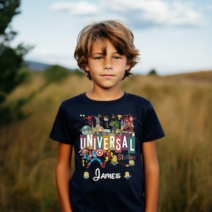 Op de afbeelding: Een marineblauw T-shirt met de tekst "UNIVERSAL Studios" en de naam "James" in witte letters. Het shirt heeft kleurrijke cartoonfiguren en logo's. De jongen draagt het T-shirt.