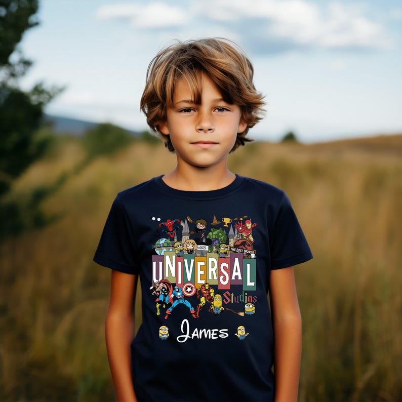 Op de afbeelding: Een marineblauw T-shirt met de tekst "UNIVERSAL Studios" en de naam "James" in witte letters. Het shirt heeft kleurrijke cartoonfiguren en logo's. De jongen draagt het T-shirt.