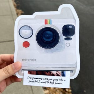 May include: A white Polaroid camera-shaped sticker with a rainbow stripe and the word "potaroid". The sticker has a photo print that reads "Every memory with you feels like a snapshot I want to keep forever."