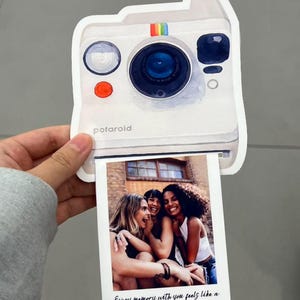 May include: A white Polaroid-style photo card with a watercolor illustration of a camera. The card features a photo of three women laughing. Text on the photo reads, "Every memory with you feels like a snapshot I want to keep forever."