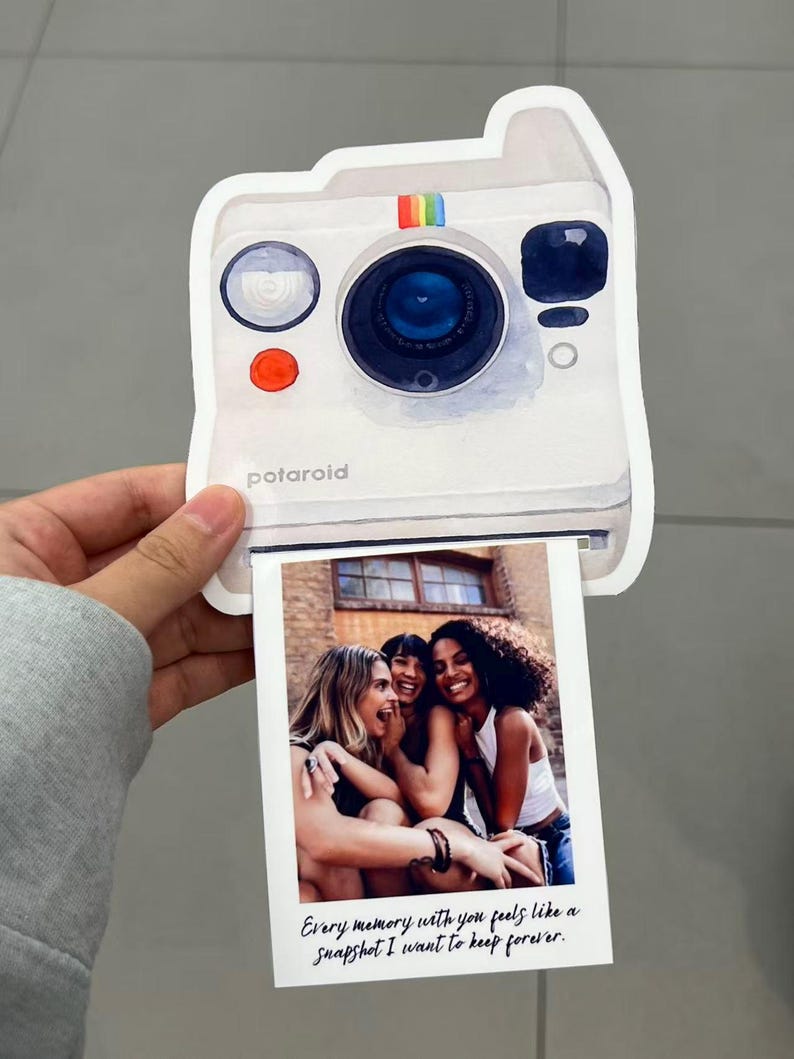 May include: A white Polaroid-style photo card with a watercolor illustration of a camera. The card features a photo of three women laughing. Text on the photo reads, "Every memory with you feels like a snapshot I want to keep forever."