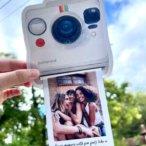 May include: A white Polaroid-style photo holder with a rainbow stripe detail and the word "polaroid" printed on the front. The photo shows three smiling women. Text on the photo reads: "Every memory with you feels like a snapshot I want to keep forever."