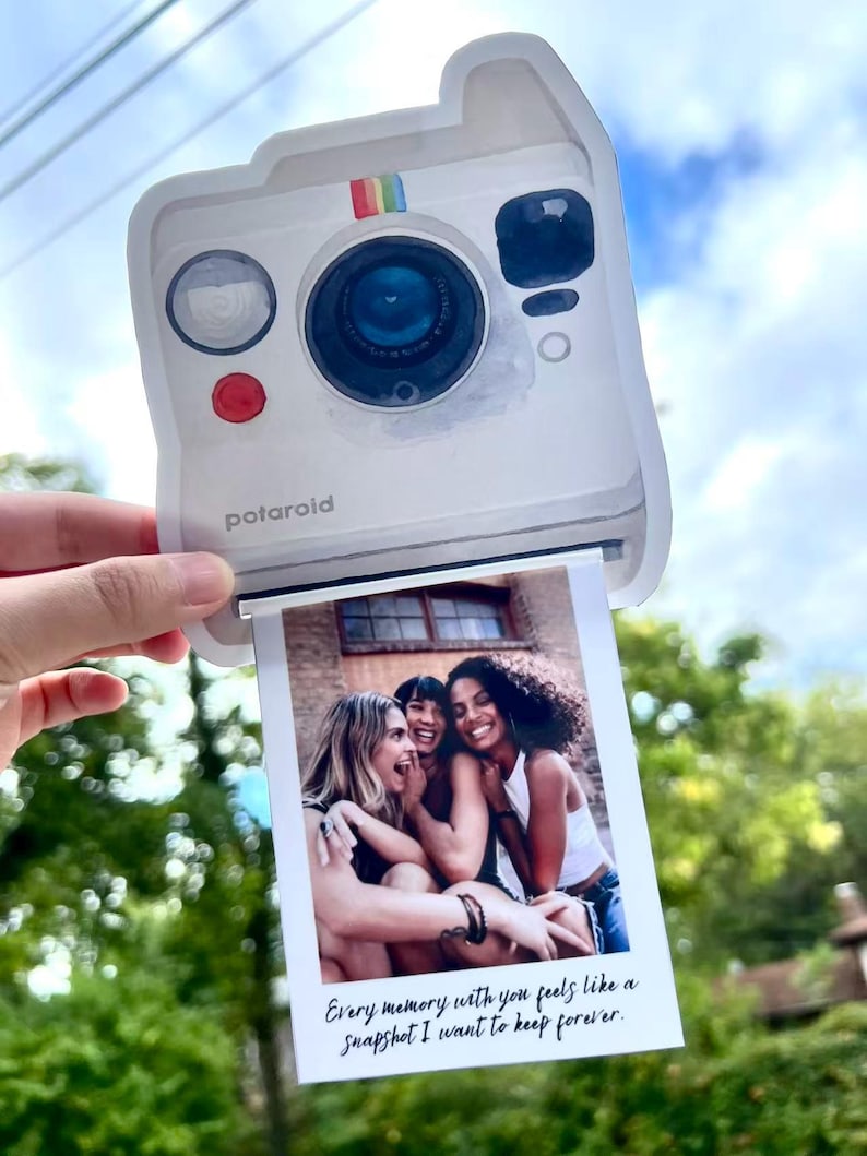 May include: A white Polaroid-style photo holder with a rainbow stripe detail and the word "polaroid" printed on the front. The photo shows three smiling women. Text on the photo reads: "Every memory with you feels like a snapshot I want to keep forever."