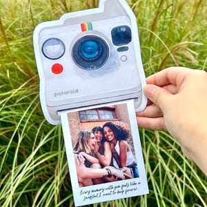 Op de afbeelding: Een witte Polaroid-stijl fotohouder met een regenboogstreepdetail en het woord "potaroid" op de voorkant. De houder toont een foto van drie lachende mensen. De foto heeft de tekst: "Every memory with you feels like a snapshot I want to keep forever."