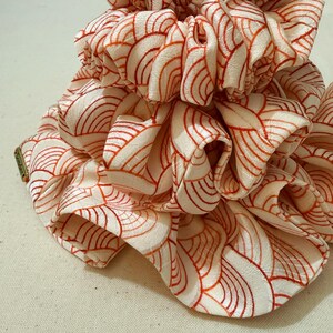 Op de afbeelding: Close-up van een stapel ivoorkleurige scrunchies met een herhalend patroon van oranje gebogen lijnen. De stof is geplooid en gerimpeld, waardoor een textuur ontstaat. De scrunchies zijn waarschijnlijk haaraccessoires.