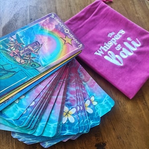 May include: A deck of tarot cards with a colorful ocean-themed design, featuring illustrations of a boat and a turtle. The cards are fanned out next to a magenta drawstring bag with the text "The Whispers of Bali."