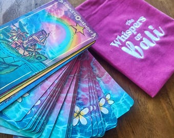 May include: A deck of tarot cards with a colorful ocean-themed design, featuring illustrations of a boat and a turtle. The cards are fanned out next to a magenta drawstring bag with the text "The Whispers of Bali."