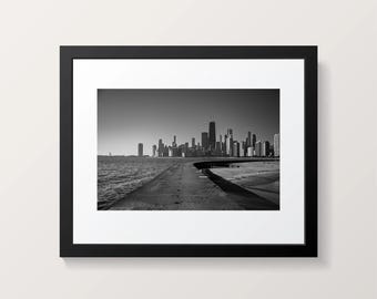 Puede incluir: Fotografía en blanco y negro enmarcada de un paisaje urbano. La imagen captura una vista frente al mar del horizonte de una ciudad, con un camino que conduce a los edificios. El cielo está despejado y el agua está en calma.