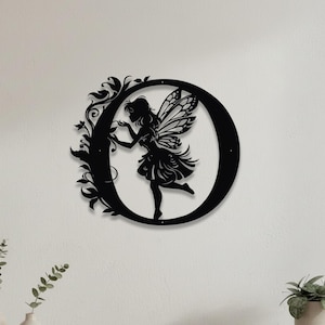 May include: A black metal wall art piece shaped like the letter O, featuring a silhouette of a fairy with wings and flowing hair. The letter is decorated with intricate vine and leaf patterns along the left side, creating a detailed cutout design.