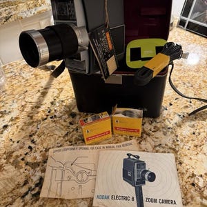 May include: A vintage Kodak Electric 8 Zoom Camera with a black and silver body, lens, and handle. The camera is in a black case with accessories, including film and manuals. The text "Kodak Electric 8 Zoom Camera" is visible.