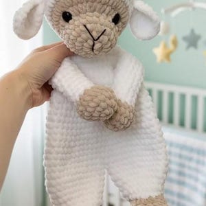 May include: A crochet lamb toy in white and beige. The lamb has a white head, body, and legs, with beige accents on the face, hands, and feet. The toy is soft and plush, with a cute, friendly expression. A lovely gift for a baby.