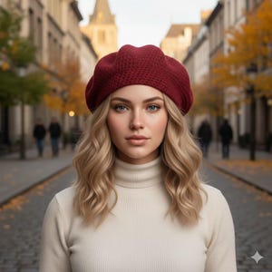 May include: A woman wearing a burgundy red beret and a cream-coloured turtleneck sweater. The beret is made of a knitted material. The background shows a cobblestone street with buildings and trees.