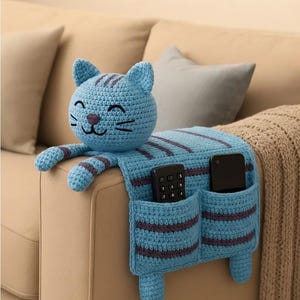 May include: A blue crochet cat-shaped couch tidy with two pockets, holding a remote and a phone. The cat has purple stripes and a smiling face. The organiser is designed to hang over the arm of a sofa.
