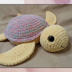 May include: A handmade crochet turtle in soft pastel colors. The turtle has a multi-colored shell in shades of pink, purple, and blue. The head and flippers are a light yellow color, with a black eye and a small smile.