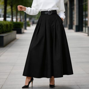 Puede incluir: Una mujer con una sudadera blanca de manga larga y una falda larga plisada negra con cinturón negro. Lleva tacones negros. El atuendo se usa al aire libre en una calle de la ciudad.