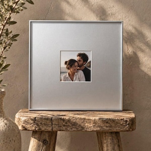 May include: A square silver picture frame featuring a wide matte border and a centred photograph of two people embracing. The frame has a clean, metallic finish and holds a small, square-shaped portrait of a couple in a close, affectionate pose.