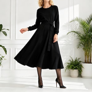 Puede incluir: Un vestido negro de manga larga con cuello redondo y cinturón a juego en la cintura. El vestido tiene una falda fluida y se combina con tacones negros. La modelo está de pie frente a una pared blanca con plantas en macetas.