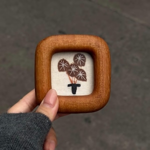 Puede incluir: Un pequeño marco cuadrado de madera que contiene una tela de color blanco roto con un diseño bordado de una planta en maceta. La planta tiene tres hojas circulares de color marrón oscuro con venas blancas cosidas en una maceta negra.