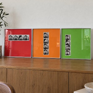 May include: Three square picture frames with thin silver borders, each featuring a vibrant solid-coloured mat in red, orange, or green. Each frame contains a vertical or horizontal black and white photo strip displaying multiple candid portraits.